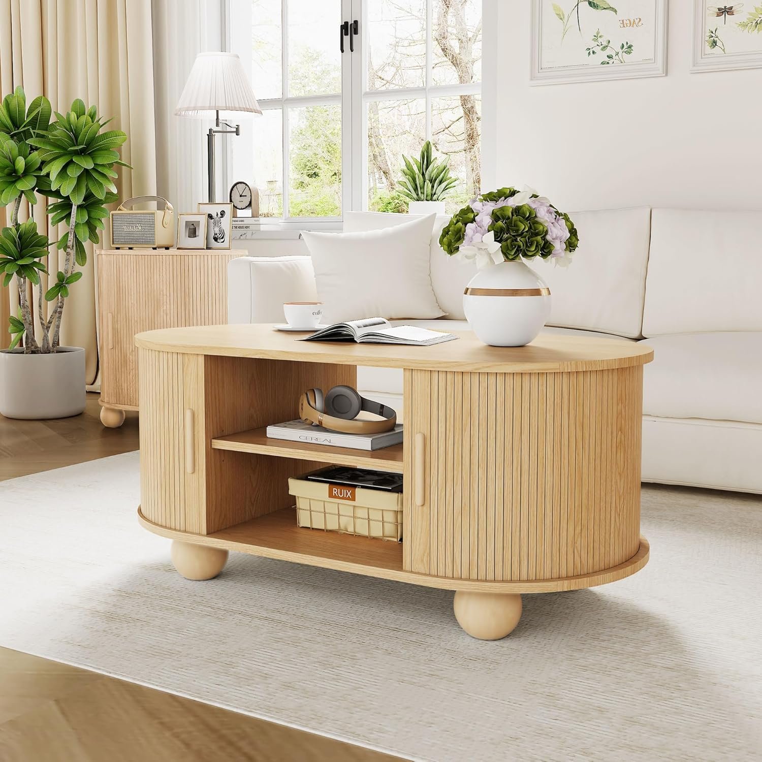 Oval fluted oak coffee table with sliding tambour doors and round ball feet, styled with a cream couch, hydrangeas, and books in a cozy living room
