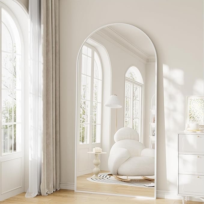 Full-length arched floor mirror with white frame, propped against a wall in a sunlit room with a boucle chair